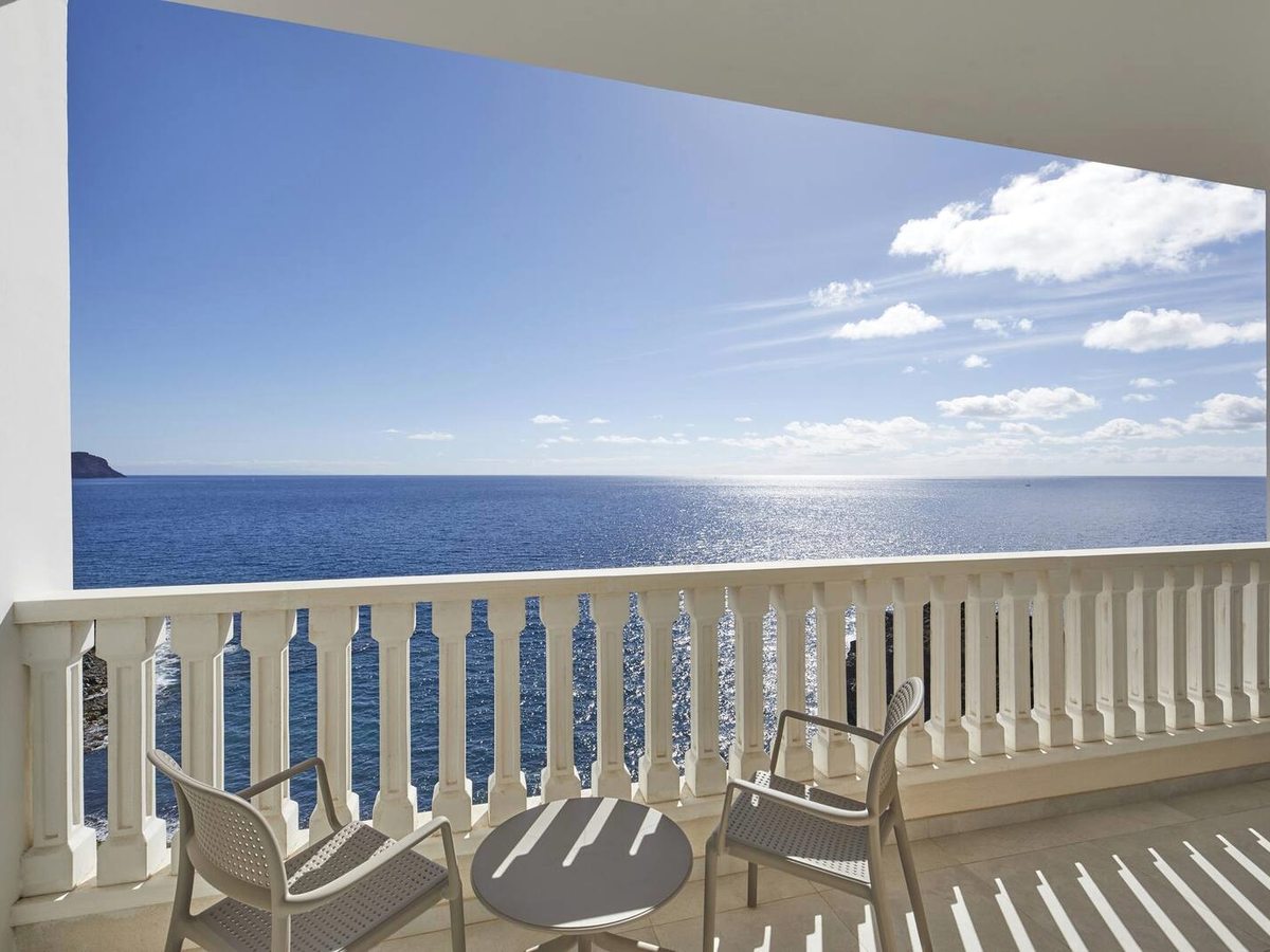 A private balcony with two chairs and a small table looking out over a calm blue Atlantic under a bright sky.