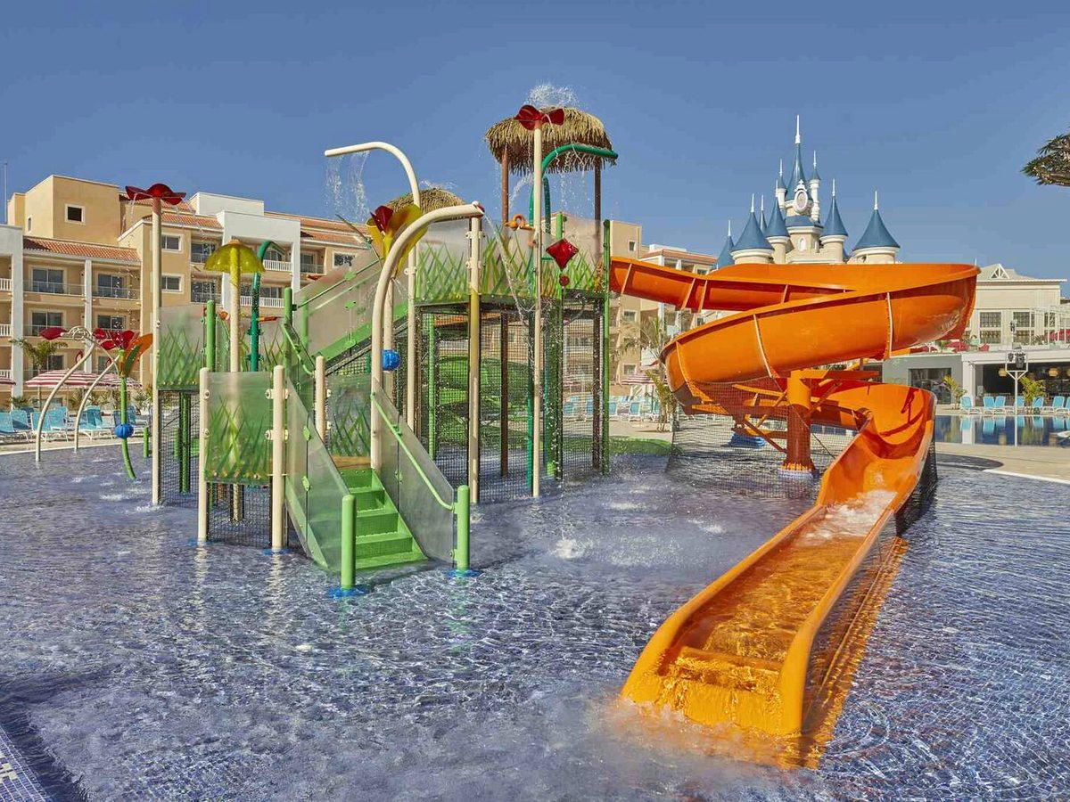 A shallow splash park with an orange spiral waterslide, green climbing structures and water-spray flowers, the castle behind.