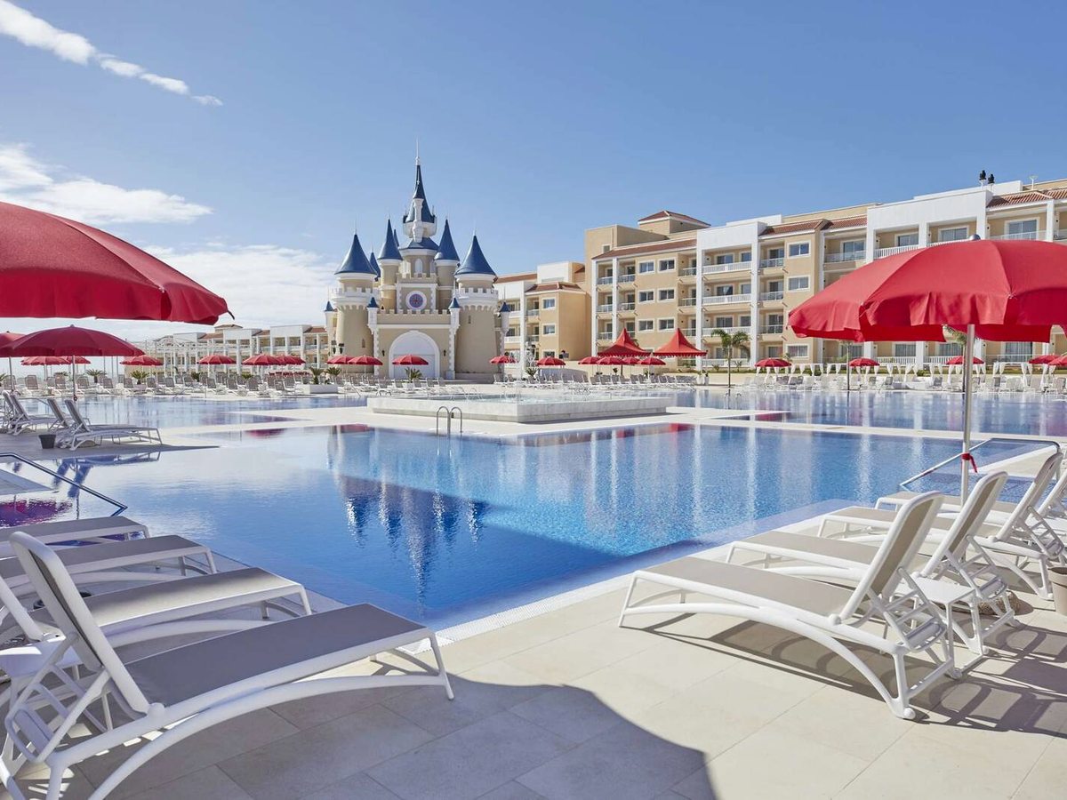 A large rectangular swimming pool lined with sun loungers and red parasols, the white castle and accommodation blocks behind.