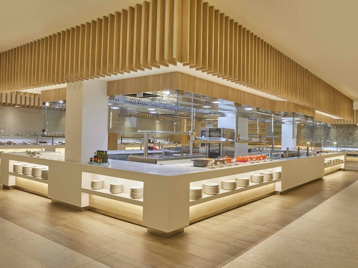A large central buffet island with glass food displays and an open kitchen in a bright, wood-ceilinged dining hall.