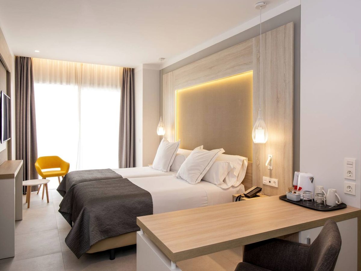 A twin-bedded room with a lit wooden headboard, a long desk with a tea tray and a yellow armchair by the curtained balcony door.