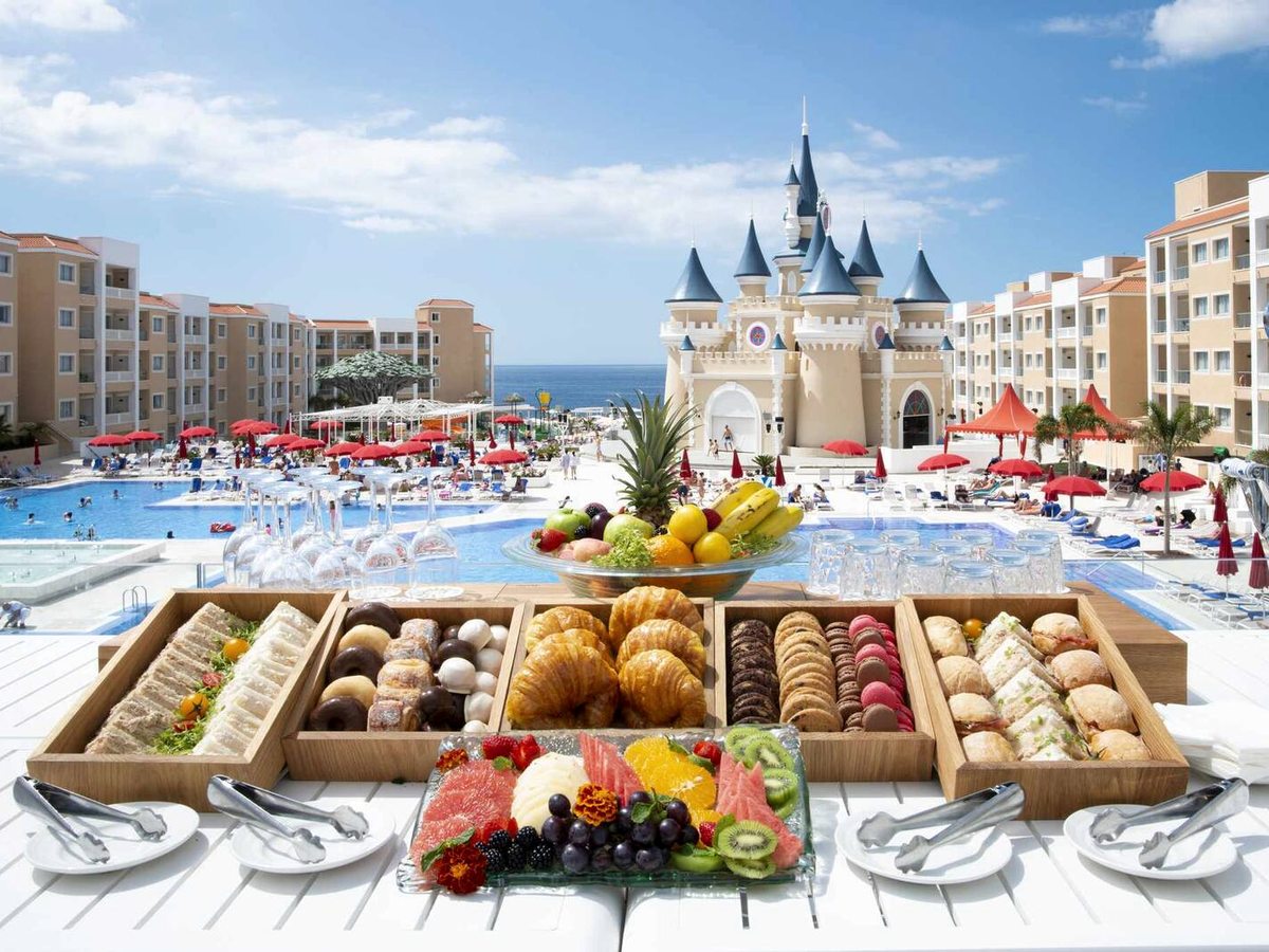 A breakfast spread of pastries, sandwiches and fresh fruit laid out on a white counter, with the castle, pool and sea behind under a blue sky.