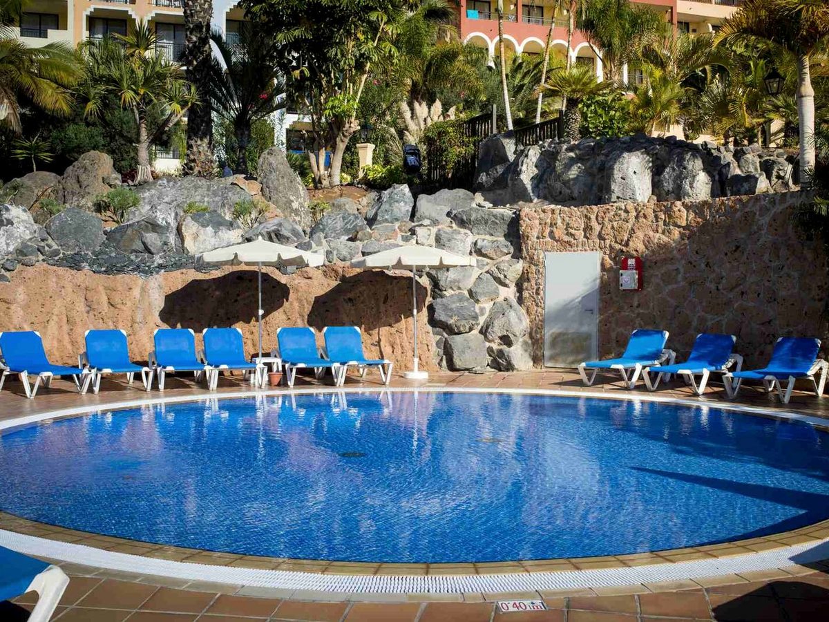 Small blue pool set against a dark volcanic-rock wall, with blue sun loungers and tropical planting around a tiled terrace.