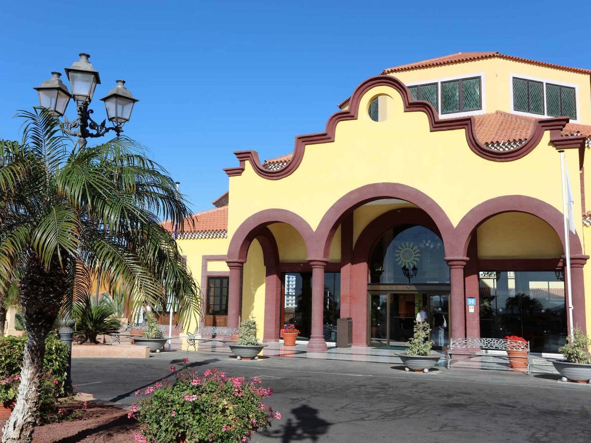 Yellow hotel entrance building with a red-trimmed arched portico, glass doors, palms and a paved forecourt under a clear blue sky.