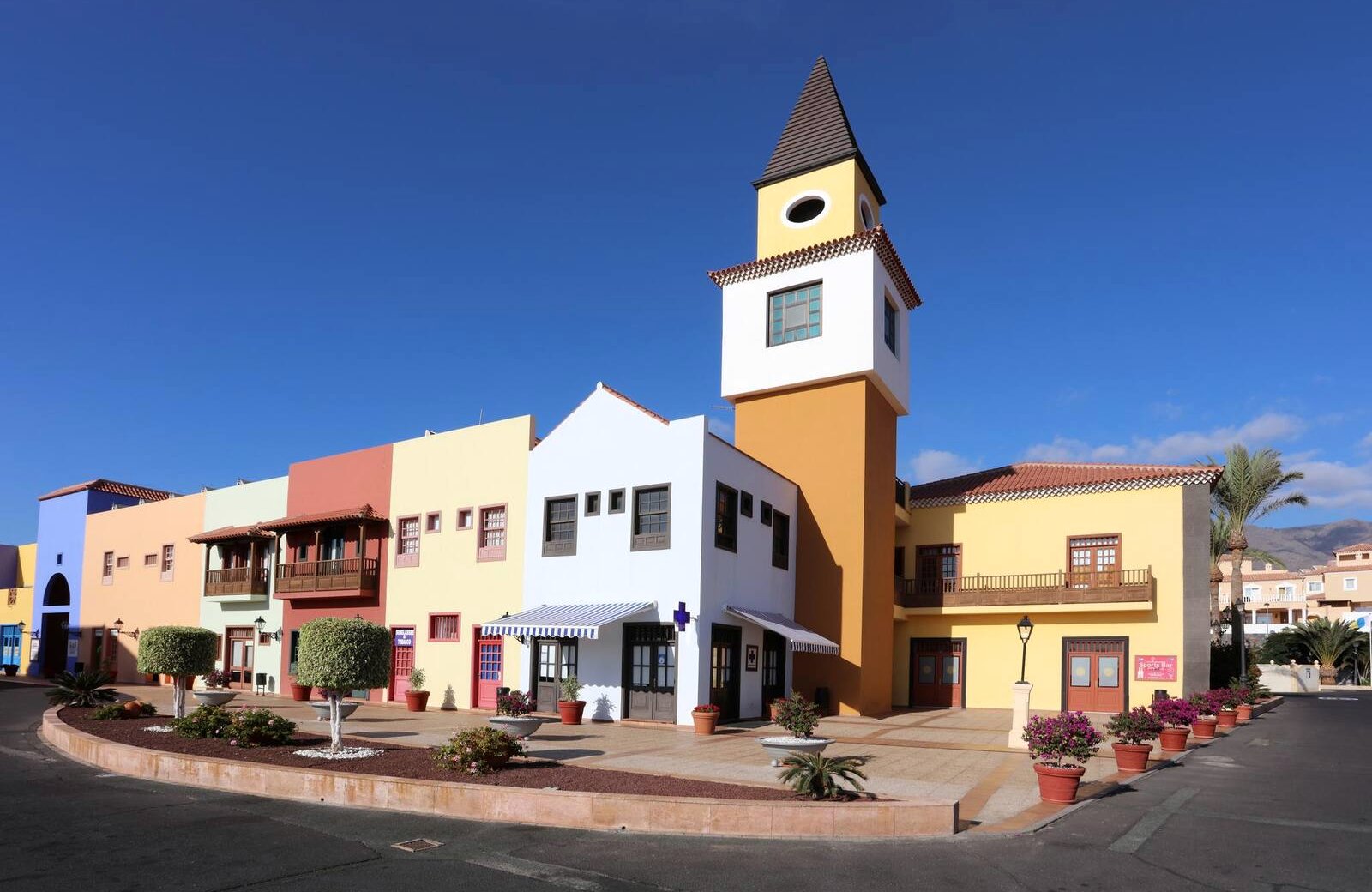 The colourful Bahía Príncipe Village square: pastel two-storey buildings with wooden balconies and a yellow clock tower around a paved plaza under a clear blue sky.