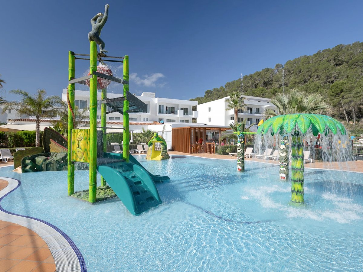 Shallow splash pool with a tall water-play tower, slides, a palm-tree fountain figure and sprinklers, backed by the pine hillside.