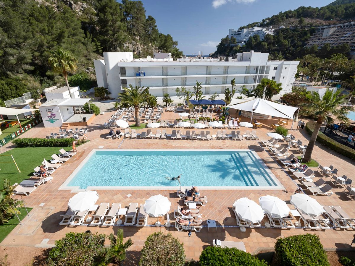 Large rectangular freshwater pool seen from above, ringed by wooden sun loungers and white parasols, with the white resort blocks and pine hillside behind.