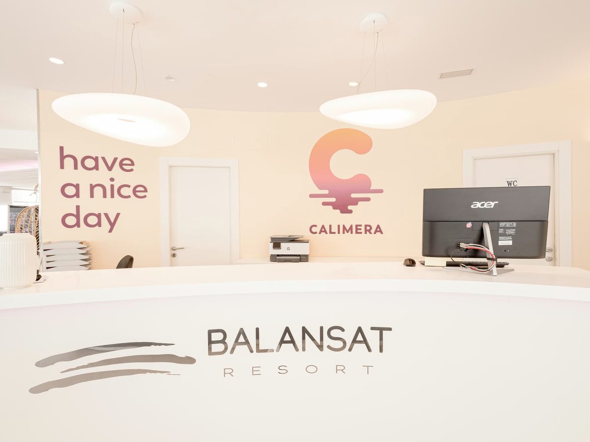 Curved reception desk with the Balansat Resort name across its front, a computer on top and a 'have a nice day' sign on the wall behind, under round ceiling lights.