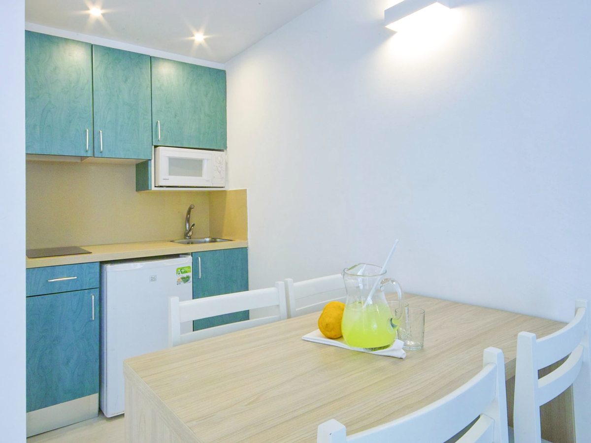 Compact kitchenette with teal units, a hob and sink, beside a pale wood dining table set with glasses and fruit.