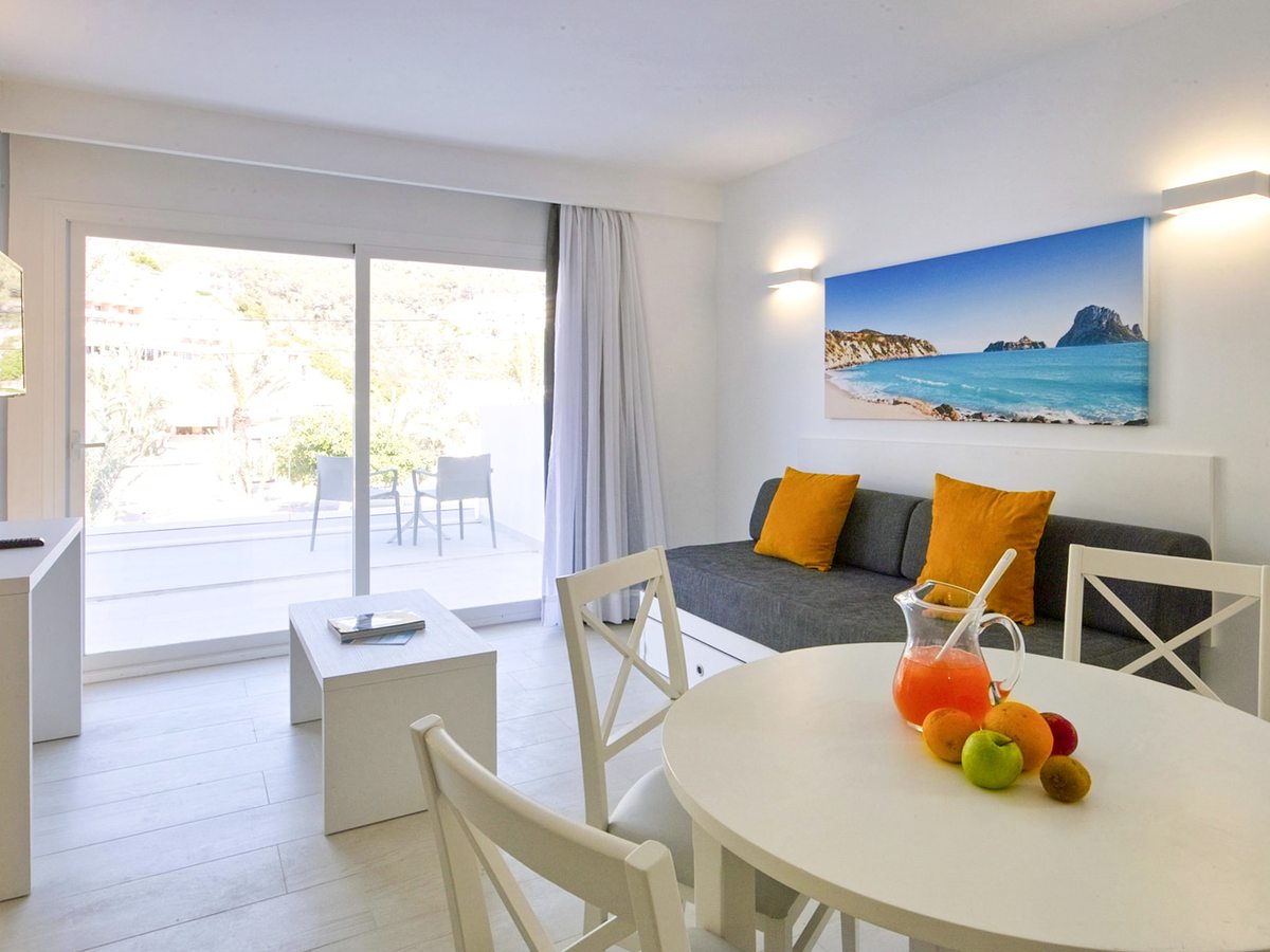 Open-plan living and dining space with a grey sofa bed, a round white dining table with a fruit bowl and doors to a private terrace.