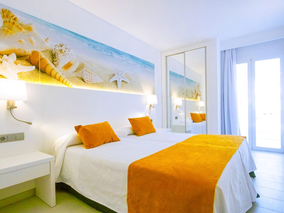 Bedroom with a white double bed, an orange runner and cushions, a driftwood-and-shell headboard artwork, bedside lamps and a mirror.