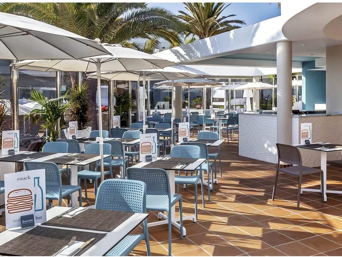 An outdoor terrace with blue chairs at set tables under large white parasols, beside palms and a servery, with menu cards on the tables.