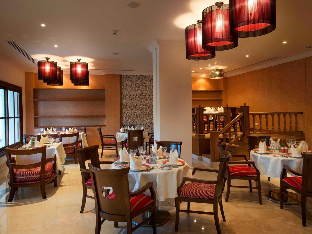 A formal restaurant with round tables under white cloths, folded napkins, wine glasses and wooden chairs with red cushions, lit by warm pendant lamps.