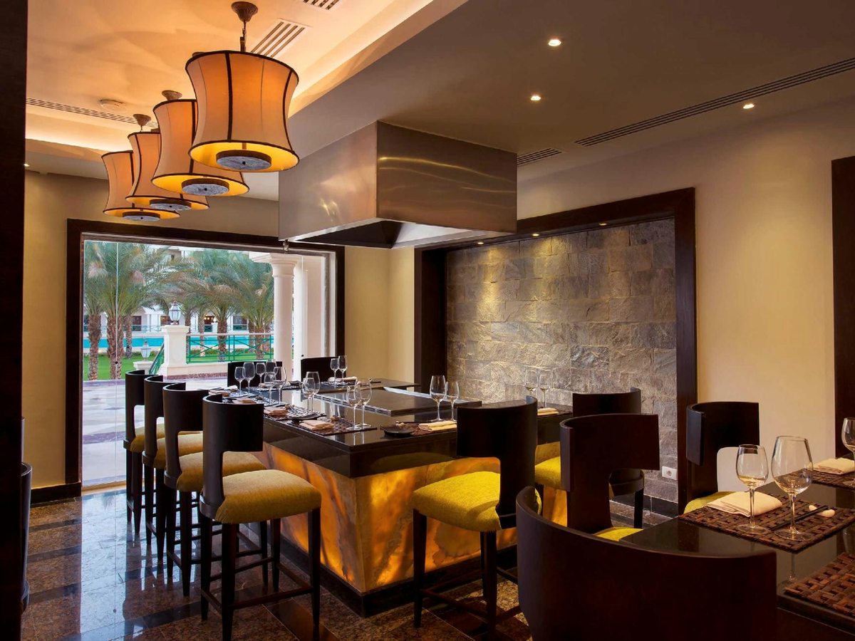 A dining counter with yellow bar stools, set tables and amber pendant lights beside an open-kitchen style counter.