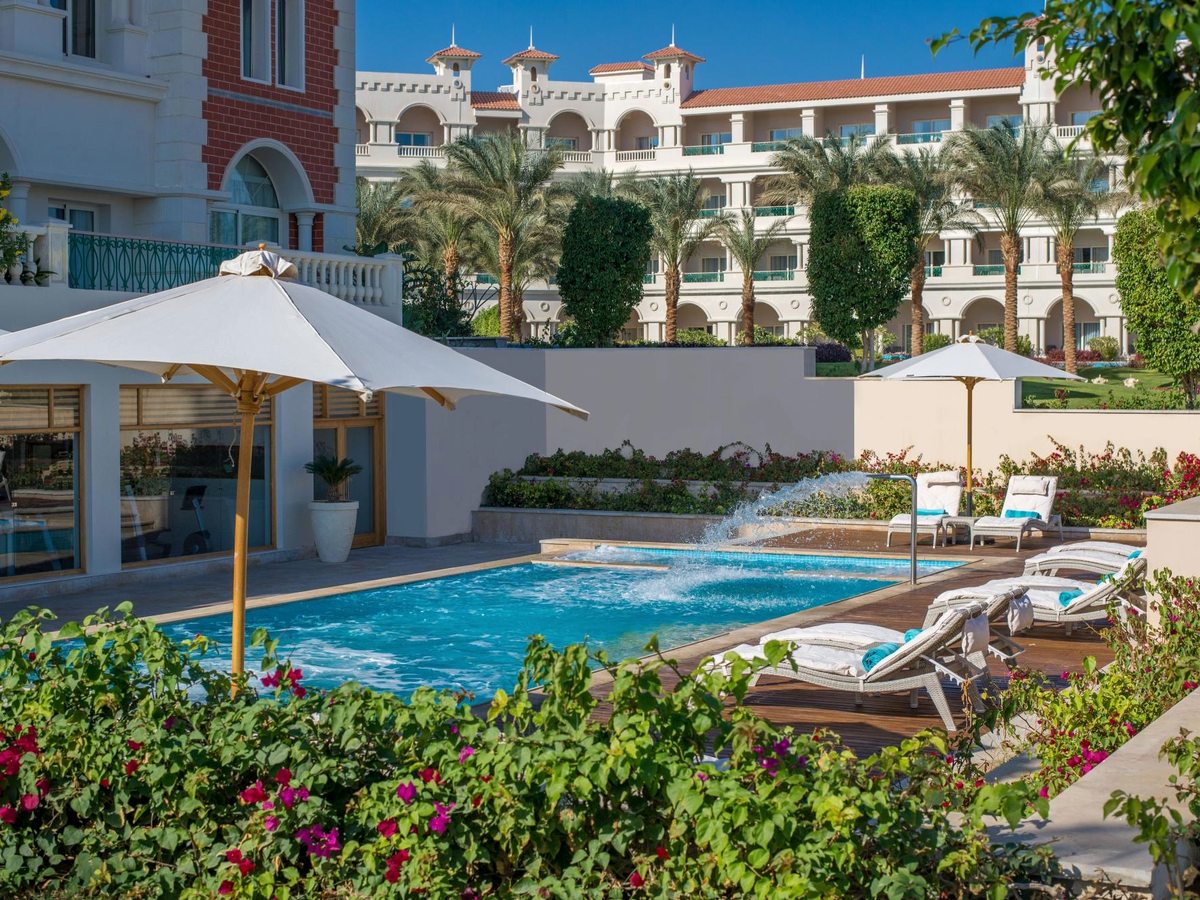 A small pool ringed by sun loungers and white parasols, with flower beds, palms and the resort's balconied wings behind.