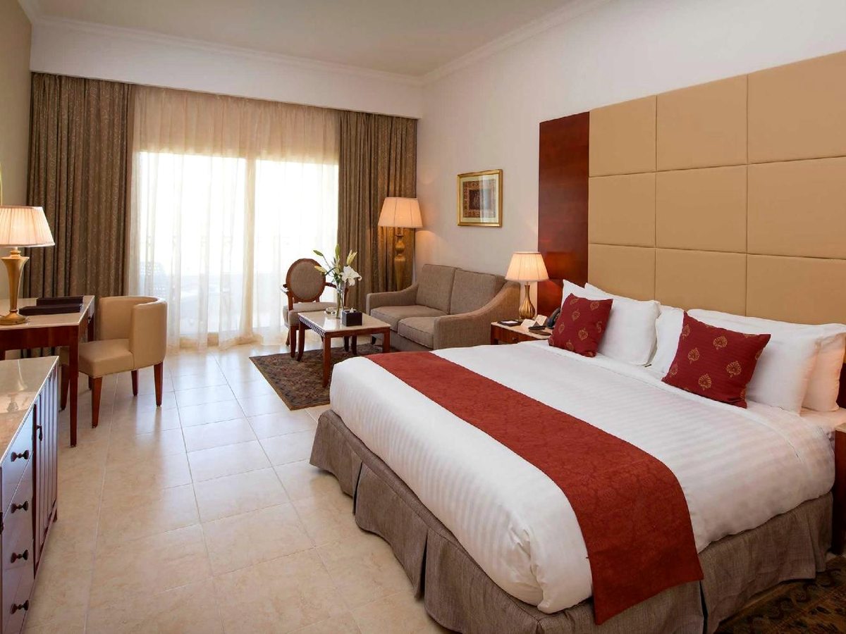 A guest room with a king bed dressed with a red runner, bedside lamps, a writing desk and tiled floor.