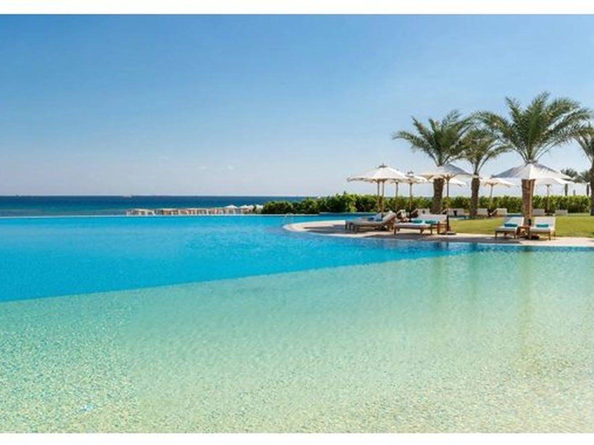 A wide turquoise pool at the water's edge with a small palm island, looking out to the sheltered bay under open sky.