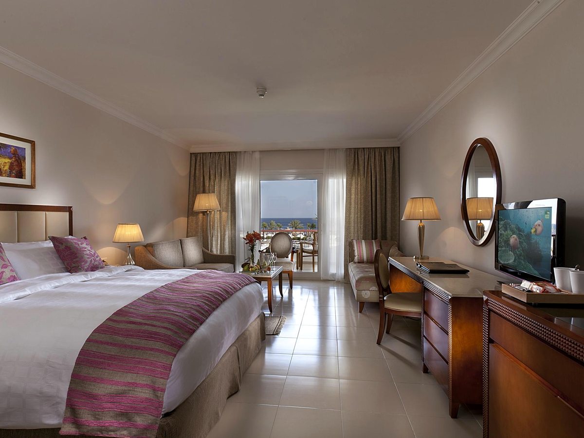 A bright guest room with a large bed, sofa seating and balcony doors opening to a sea view.