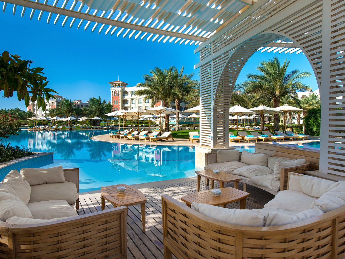 A pool deck with a white slatted pergola, wicker loungers and cushioned seating under sun umbrellas, palms and the resort facade behind.