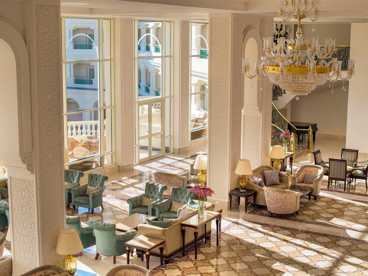 A double-height lobby lounge with tall arched windows, a crystal chandelier, marble floor and clusters of sofas and armchairs.