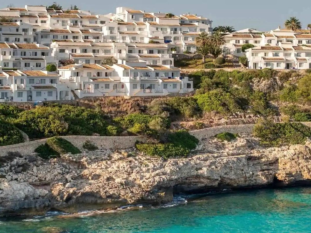 Tiered white apartment blocks with tiled roofs on a green and rocky cliffside above a turquoise cove.