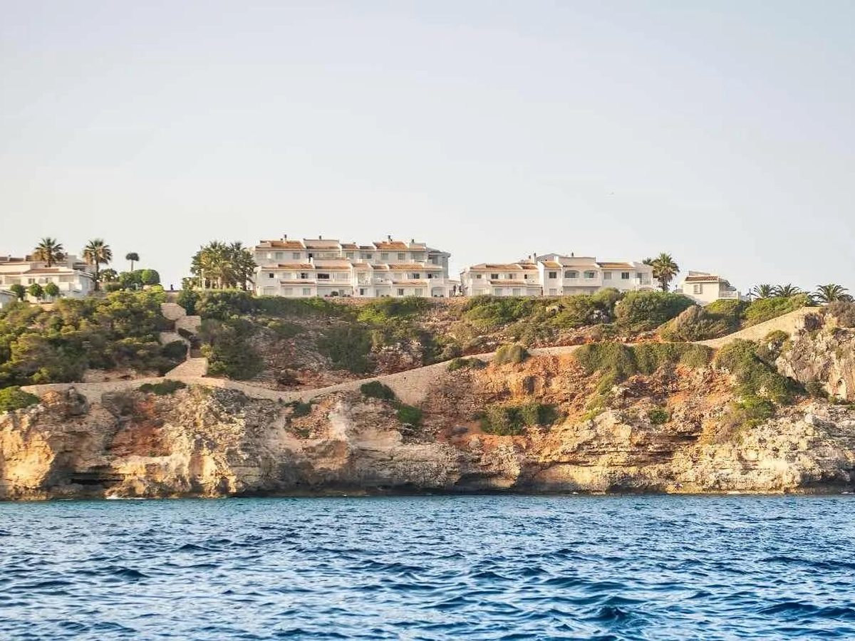 White resort buildings lining the top of a rocky cliff above deep-blue sea, viewed from out on the water.