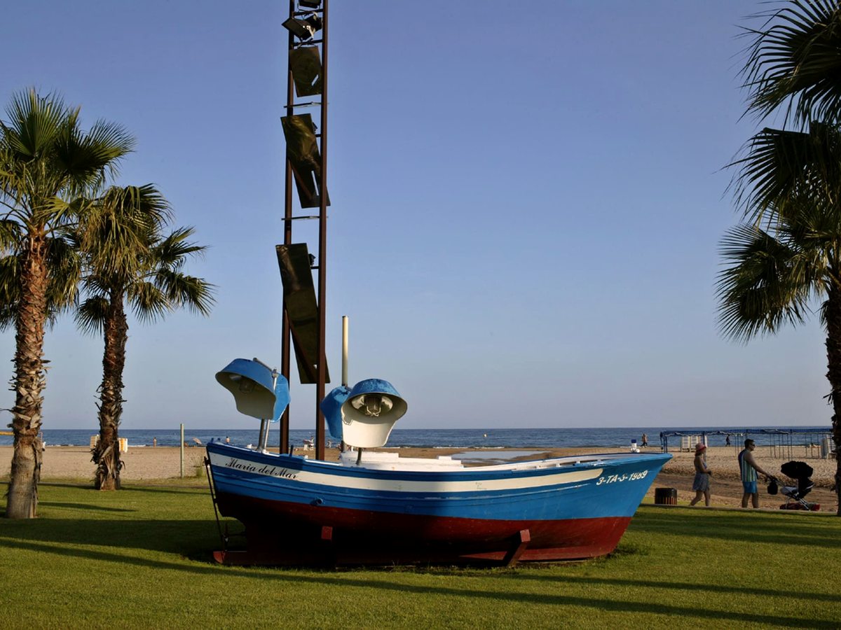 A blue, white and red wooden fishing boat mounted on grass between palms, the beach and sea behind