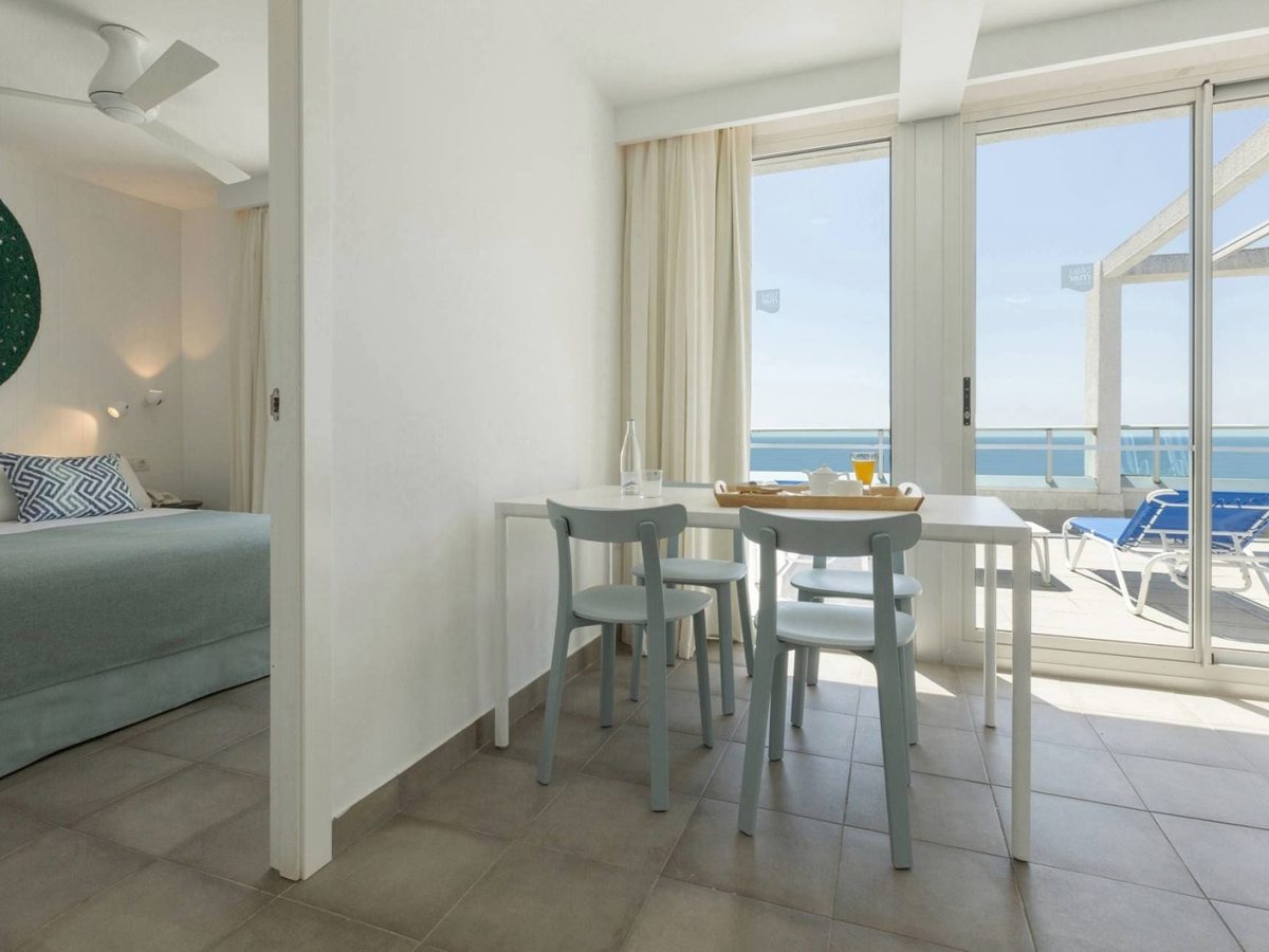 A suite dining table and pale blue chairs by sliding doors onto a terrace with sunloungers and open sea views, the bedroom to the left