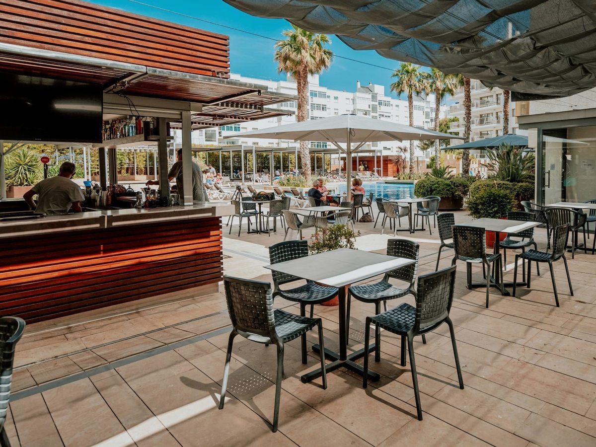 An open-air bar with a barman, wooden decking, tables and parasols beside the pool, the hotel behind
