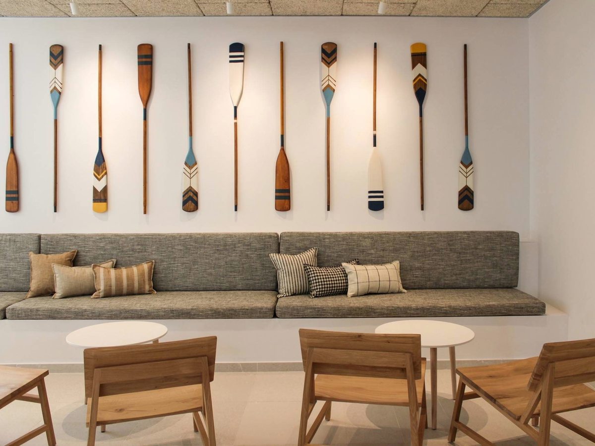 A long grey banquette below a row of painted wooden oars, with wooden lounge chairs and small round tables