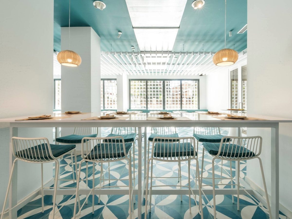 A bright bar room with white high stools at a long counter, turquoise patterned floor tiles and rattan pendant lights