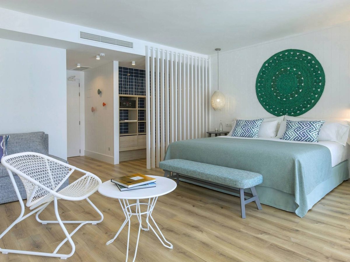 A suite with a double bed under a green woven headboard, a grey sofa, white wire chairs and a slatted divider by the kitchenette