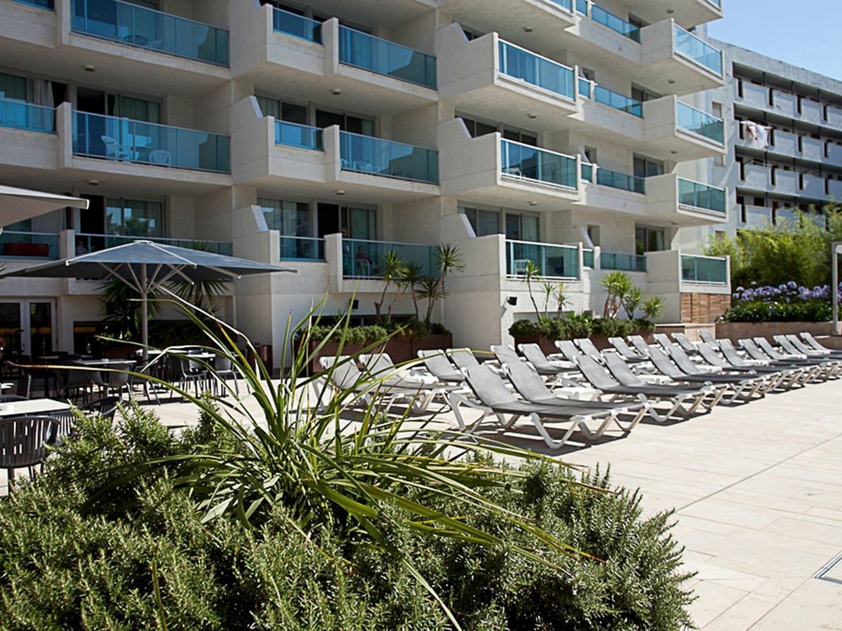 Rows of grey sunloungers on a paved terrace beside the hotel, with a parasol and planted borders