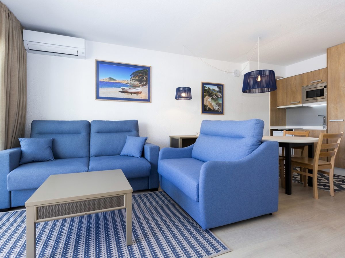 A living room with two blue sofas, framed coastal prints, a wooden kitchenette and a dining table