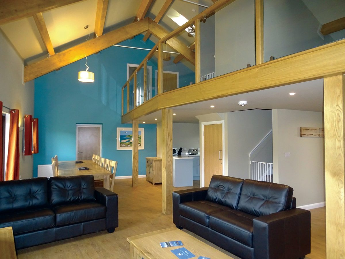 An open-plan lodge living room with a double-height ceiling, a teal feature wall, exposed timber beams, black leather sofas and a mezzanine balcony above.