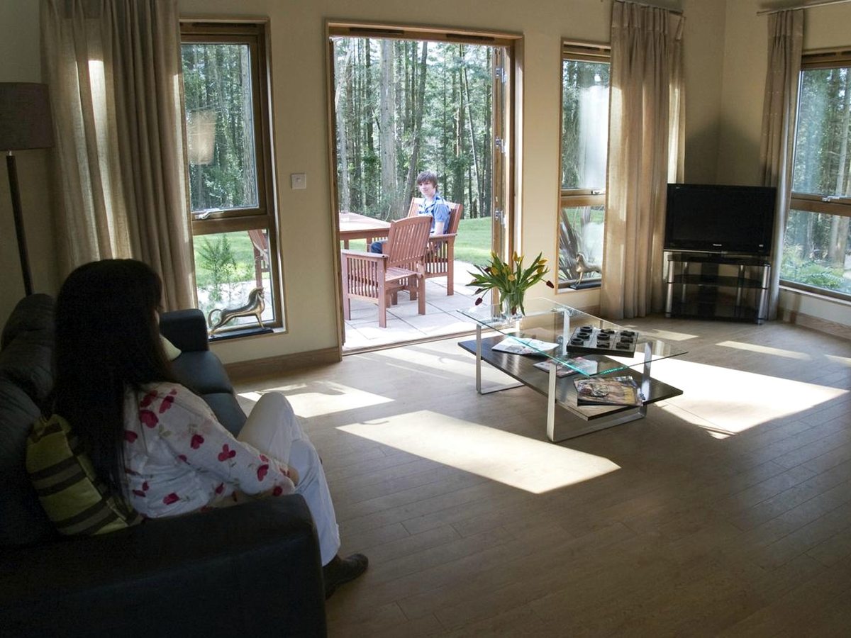 A bright lodge living room with armchairs and a coffee table, tall windows and patio doors opening onto a woodland deck with outdoor chairs.
