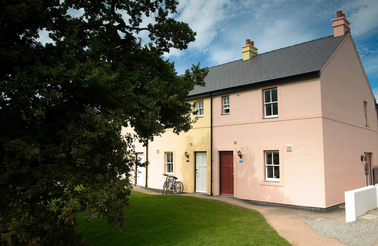 Bluestone National Park Resort: colourful pastel-rendered Village cottages with slate roofs, a bike leaning against one and a lawn with a mature oak tree.
