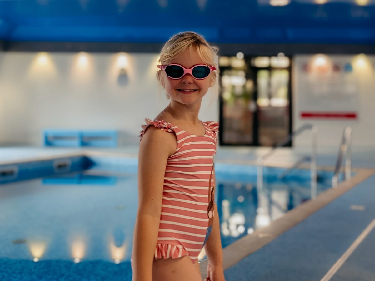 A child in a striped swimsuit and sunglasses standing at the tiled edge of Bosinver's blue indoor pool.
