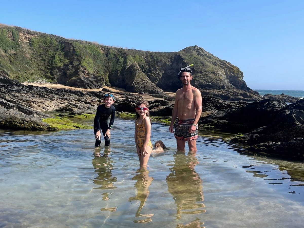 A family paddling in a shallow natural rock pool below rugged sea cliffs on the Cornish coast.