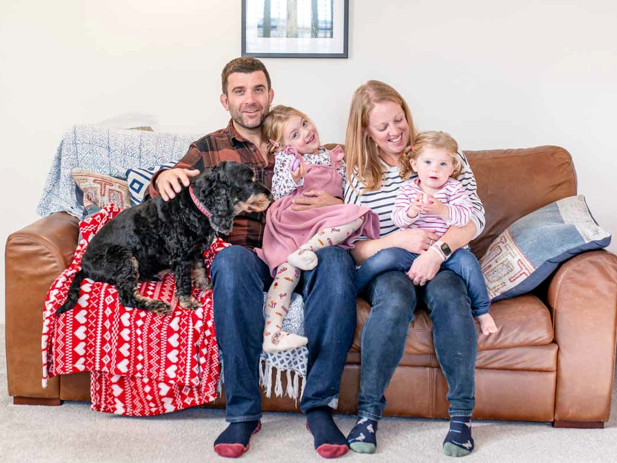 A family with two young children and a black dog on a brown leather sofa with patterned cushions and blankets in a Bosinver cottage sitting room.