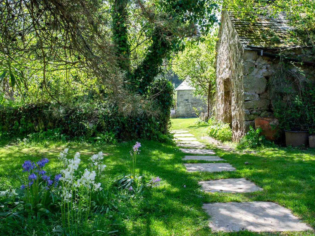 A stepping-stone path across a sunny lawn with bluebells and a stone outbuilding in a Bosinver cottage garden.