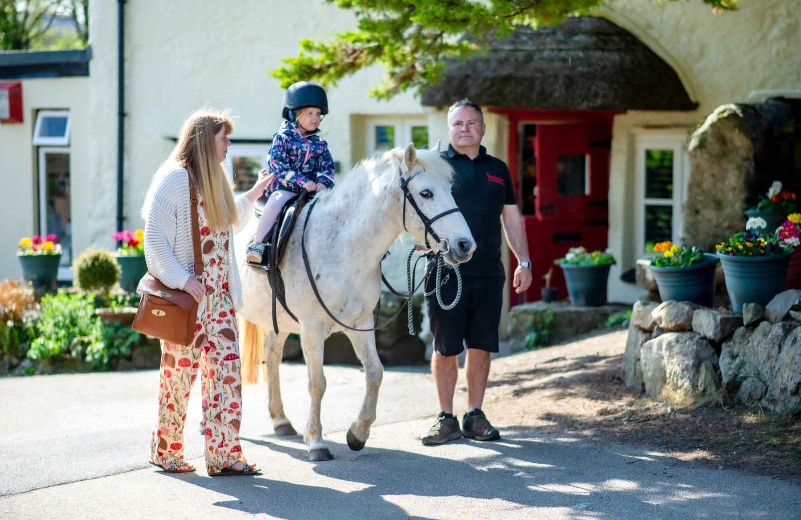 A toddler in a riding hat on a white pony, led past a whitewashed thatched cottage with a red door at Bosinver Farm Cottages.
