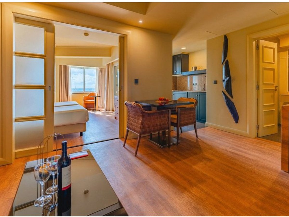 Open-plan suite with a wicker dining table, a kitchenette along the wall and sliding doors through to a sea-facing bedroom.