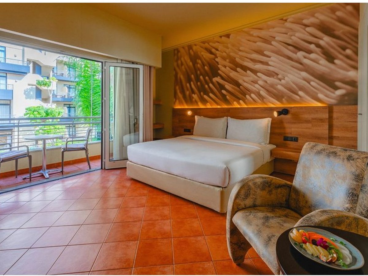 Hotel room with a double bed, a patterned armchair and a balcony overlooking the resort buildings and greenery.