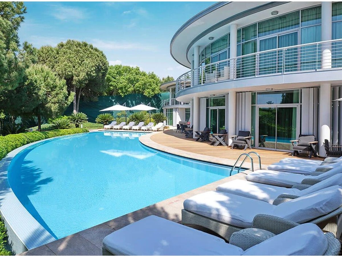 A pool beside a curved resort building, with cushioned double loungers and parasols lined along the tiled surround.