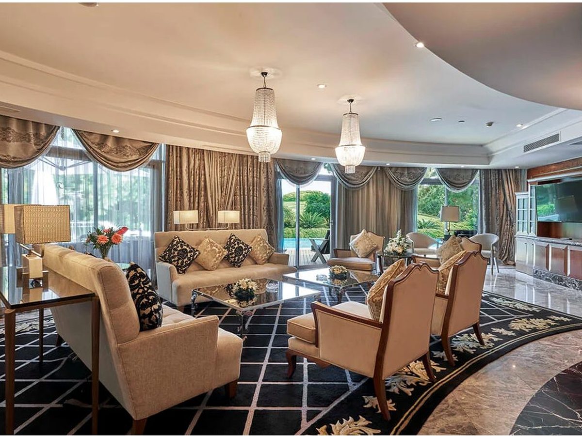 A suite living room with cream sofas and armchairs, a patterned rug and a chandelier, floor-to-ceiling curtained windows to the garden.