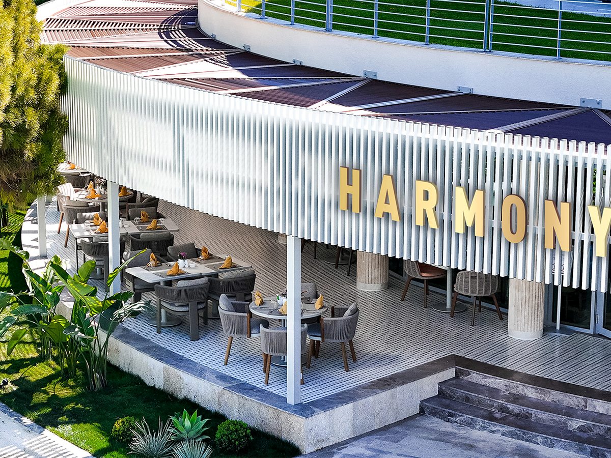 The Harmony restaurant exterior with gold HARMONY lettering on a white curved canopy above a covered dining terrace, steps and garden planting.