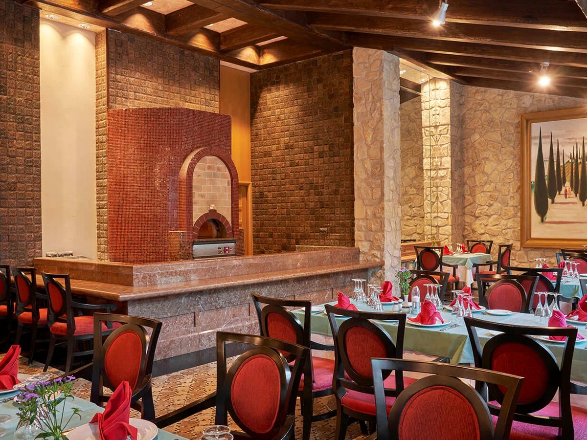The rustic dining room of Timo, with a red-tiled wood-fired brick oven, stone walls, timber-beamed ceiling and red-upholstered chairs at set tables.