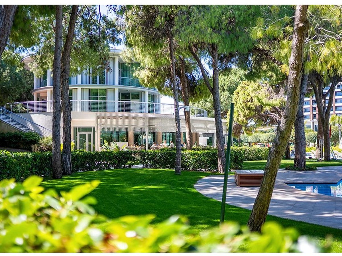 Landscaped resort gardens with clipped lawns and a curved path winding between tall stone pines, a low white resort building beyond.