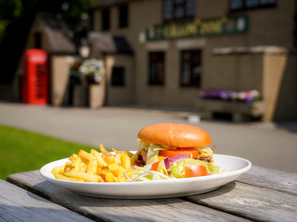 A burger in a bun with a bowl of chips and salad on a wooden picnic table, the Inn behind.